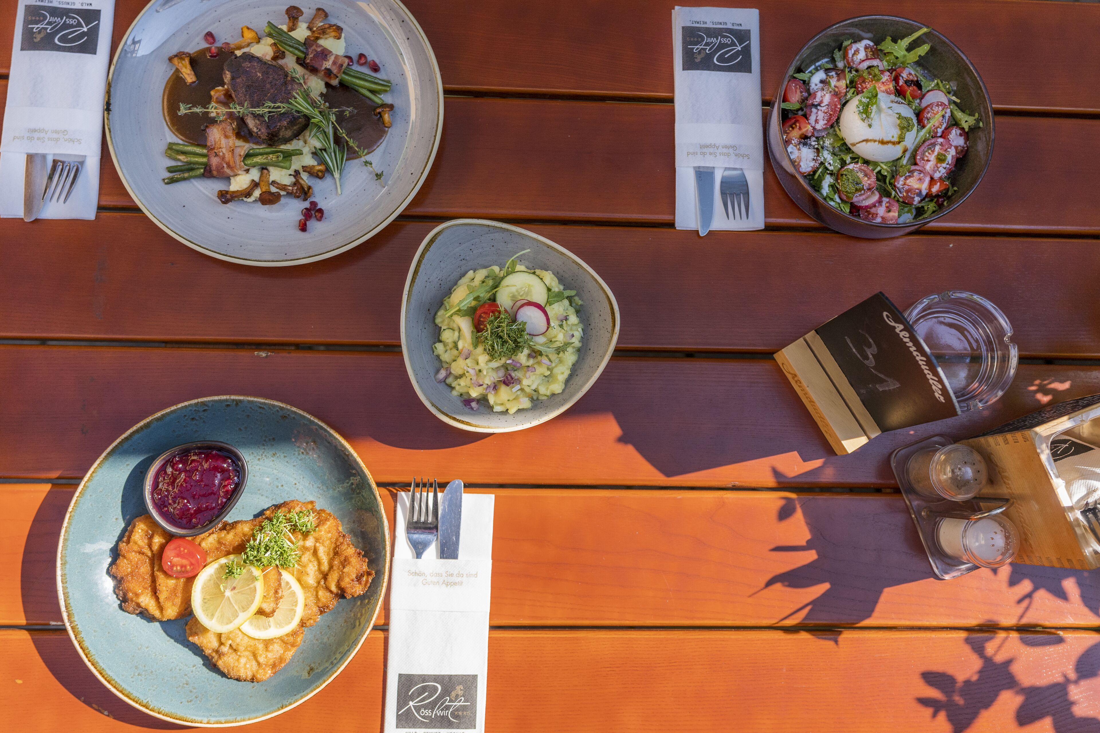 Una mesa de madera con varios platos, entre ellos una ensalada, un guiso de carne y ensalada de patatas, junto con salsas y cubiertos.
