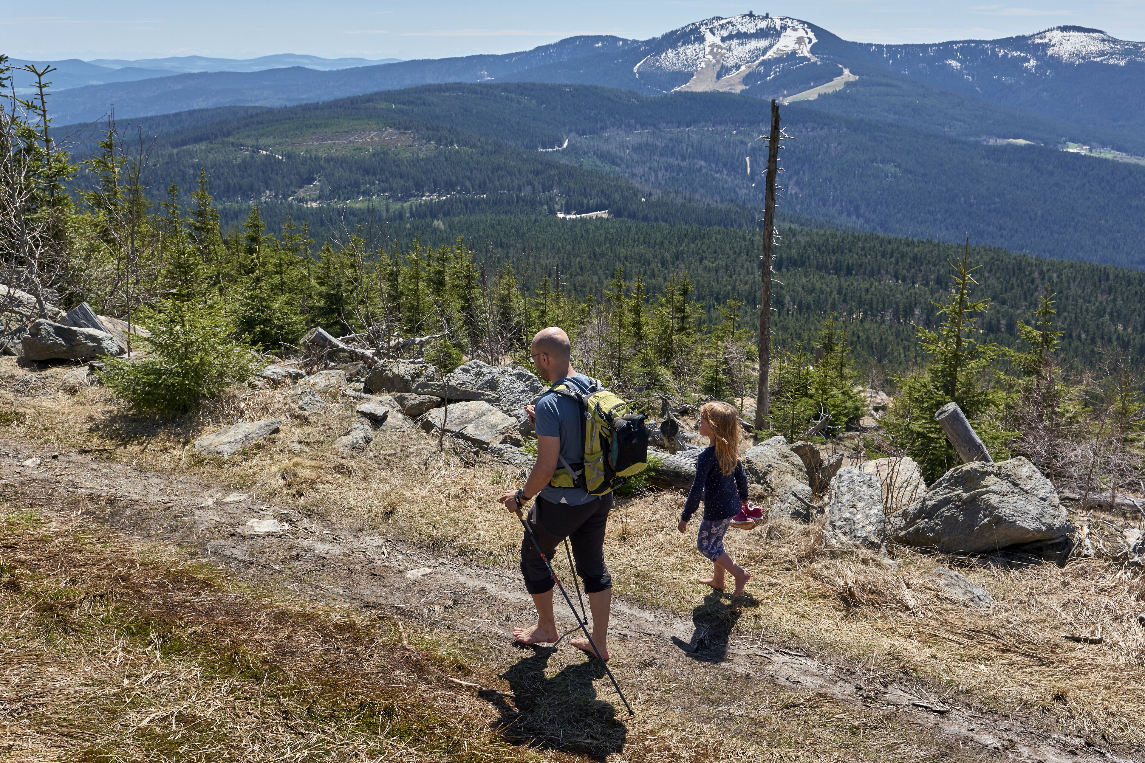 Un excursionista con una mochila y un niño caminando sobre un sendero rocoso, con un paisaje montañoso al fondo.