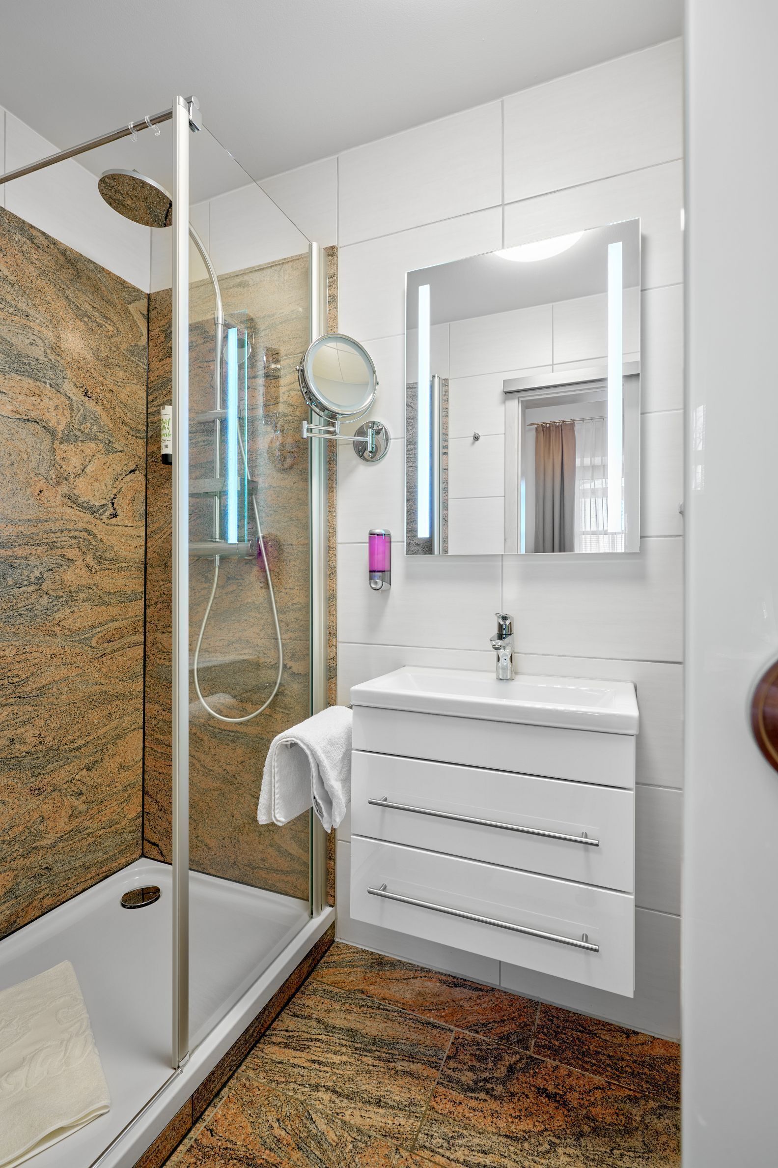 Un baño moderno con ducha envuelta en vidrio, un lavamanos blanco con espejo y piso de baldosas de mármol.