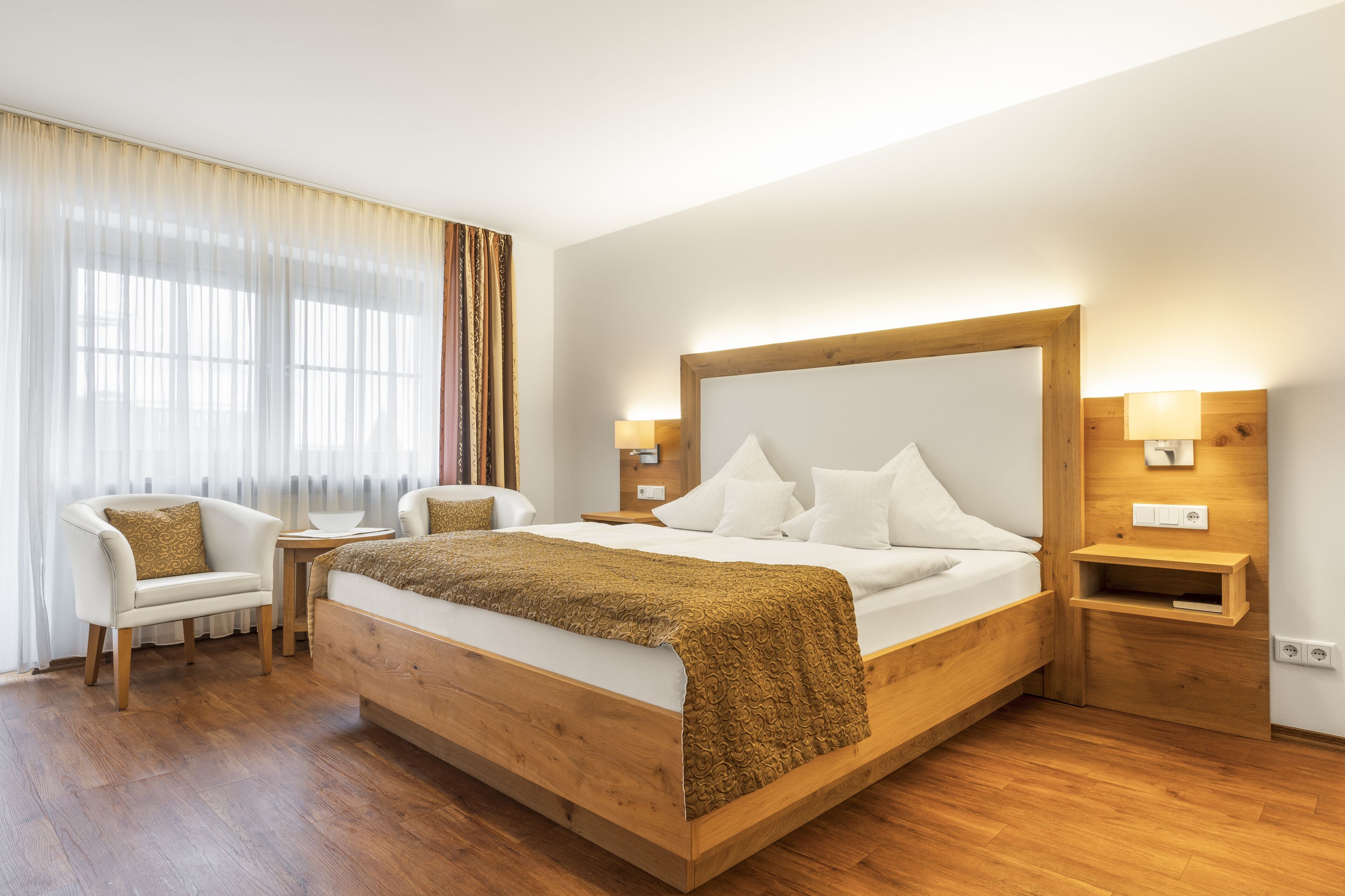 A modern hotel room with a wooden bed, white linens, a small bedside table, a chair, and a large window with sheer curtains.