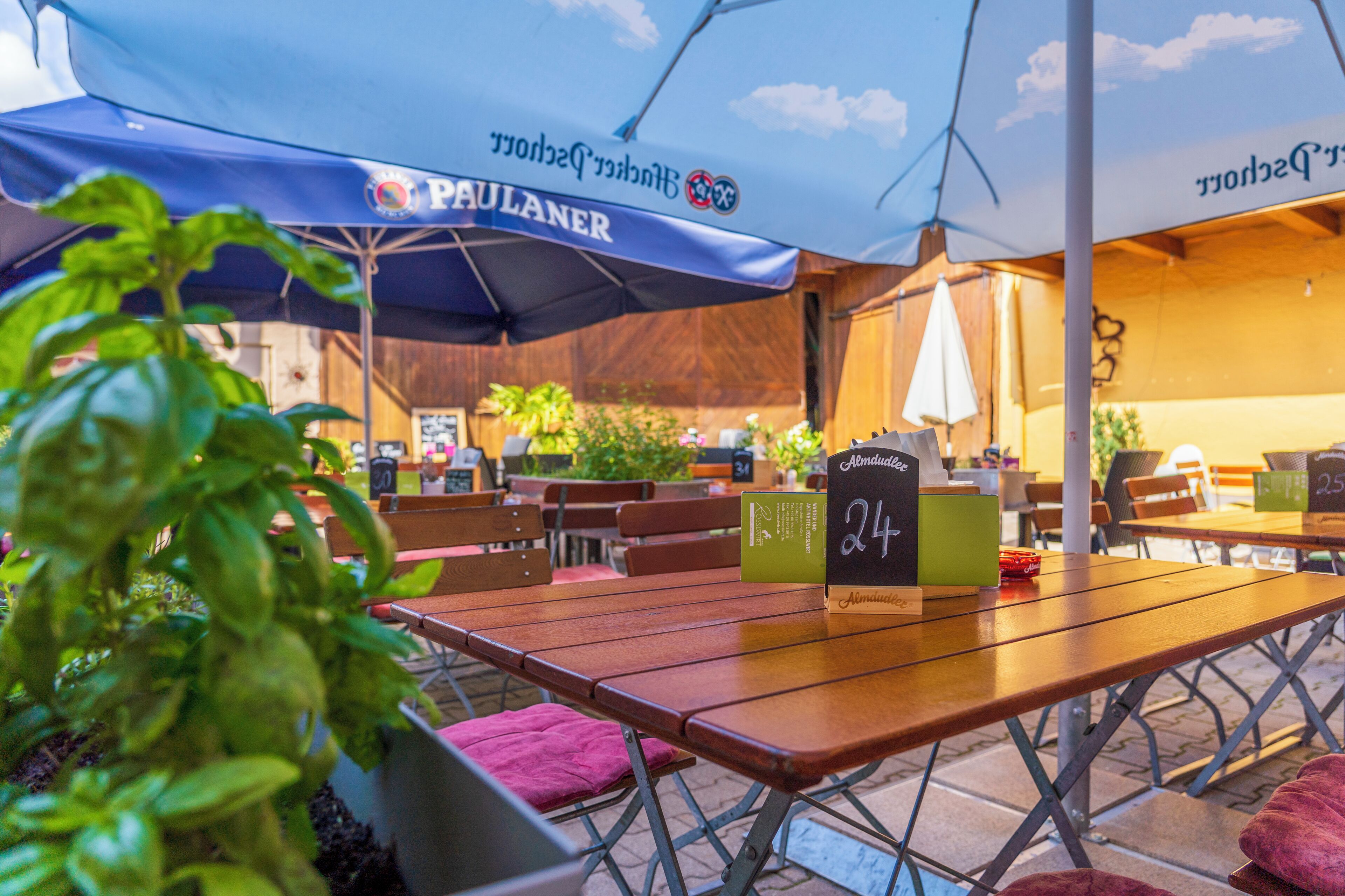 An outdoor café with wooden tables and blue umbrellas labeled 'PAULANER.'