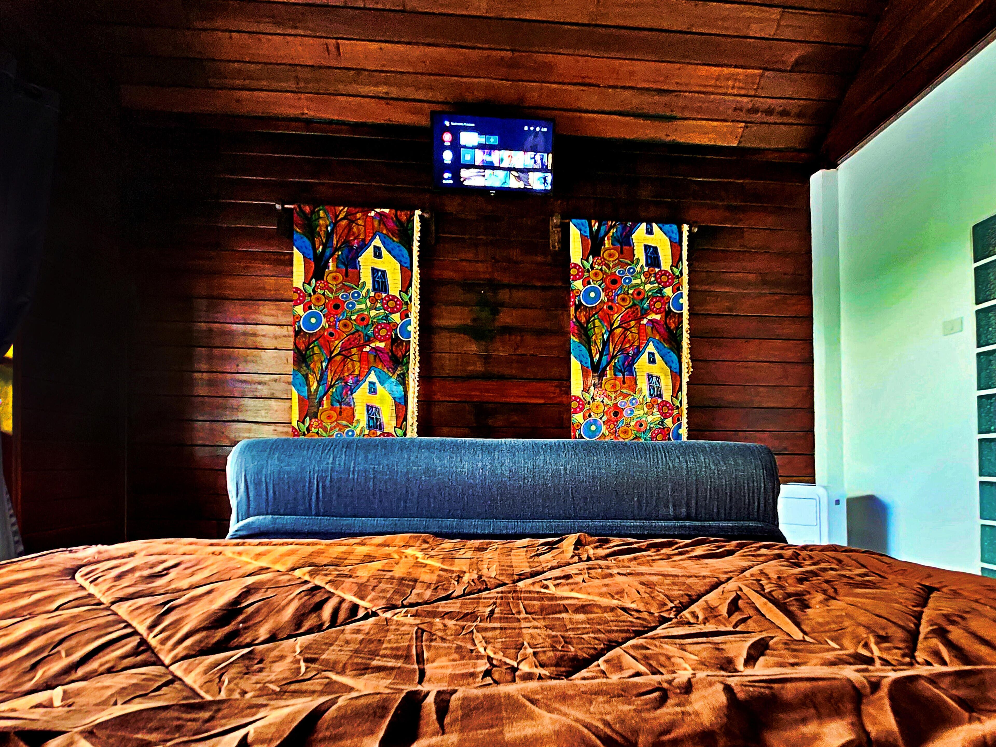 Quarto com cama, paredes de madeira, televisão na parede e cortinas coloridas.