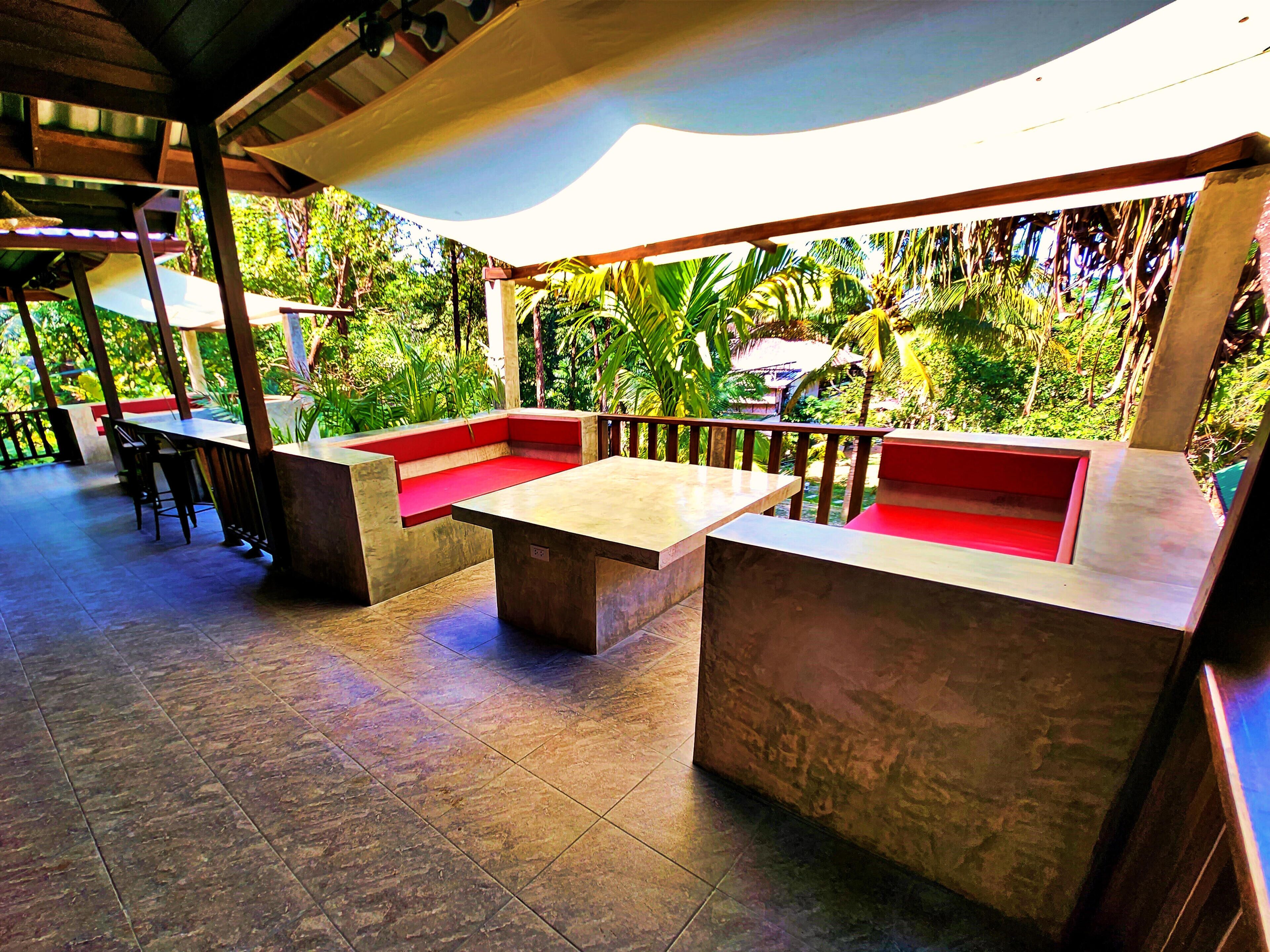 An outdoor seating area with stone tables and red cushioned benches, surrounded by lush greenery.