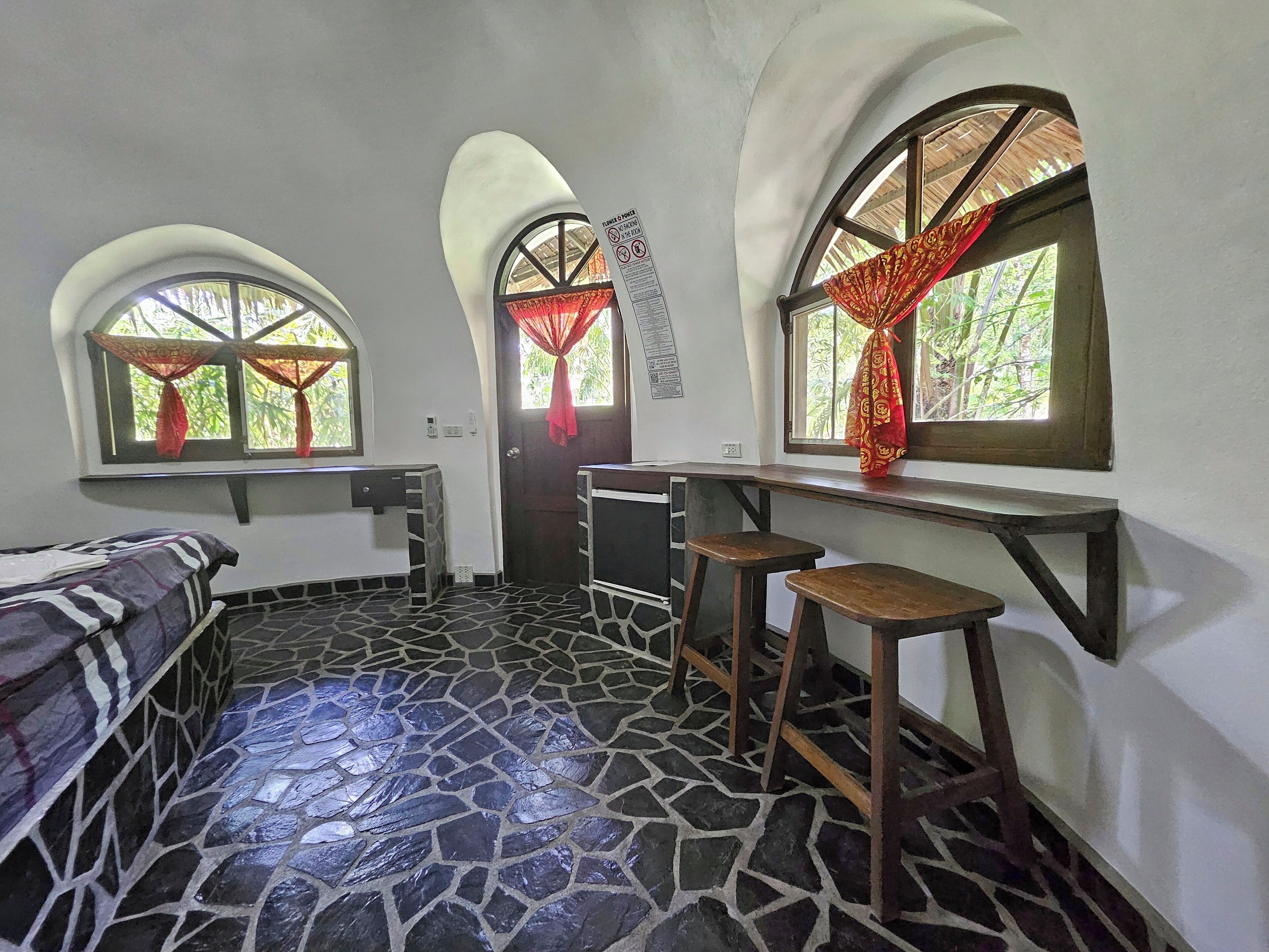 A room with a bed, a desk, two stools, and a window with a red curtain.