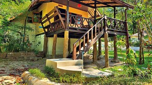 A wooden structure with a balcony and stairs leading to a yellow building surrounded by greenery. - Flower Power Village (Ranong)