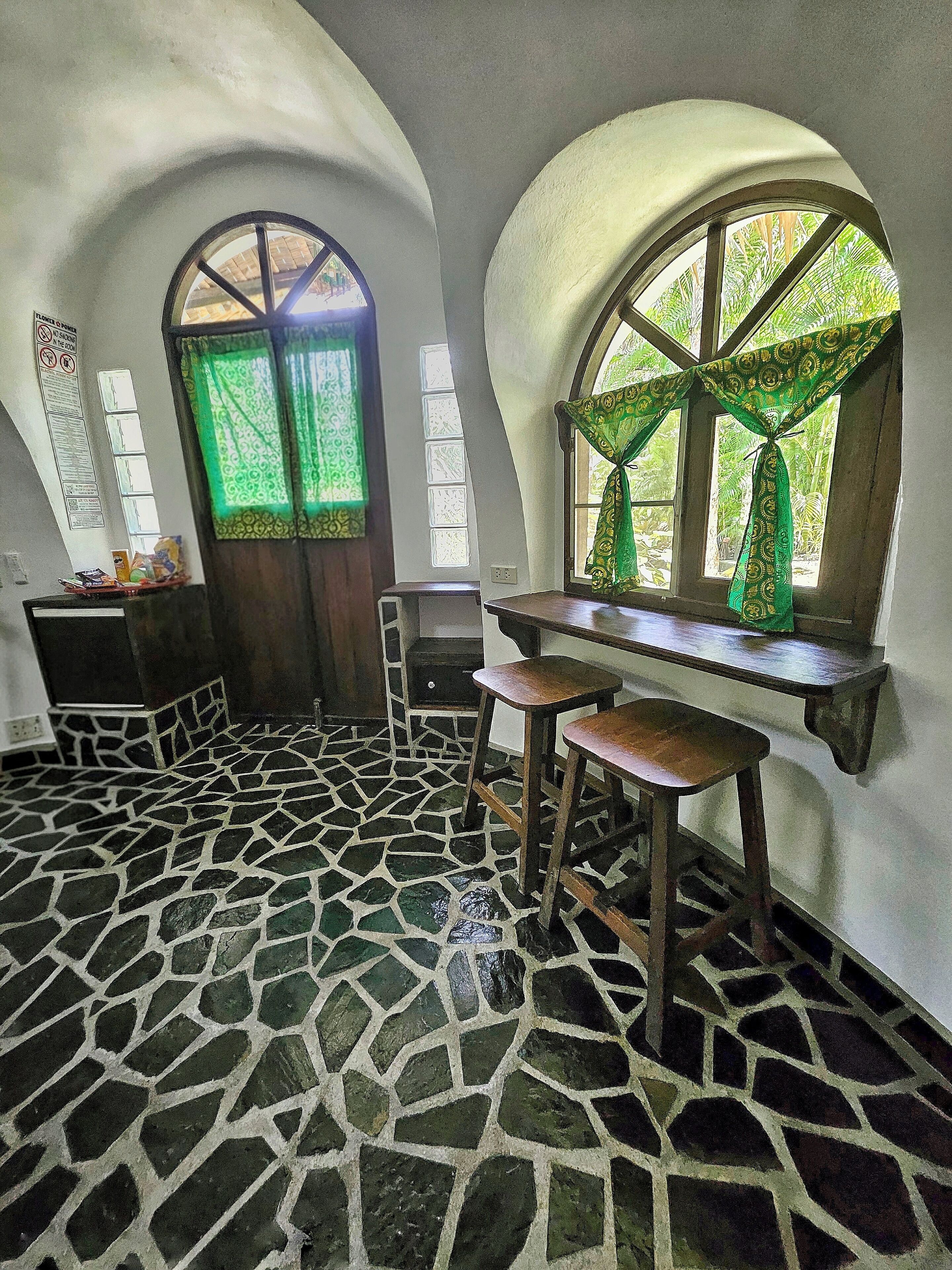 A room with a stone-patterned floor, a wooden bench, a desk, and a window with green curtains.