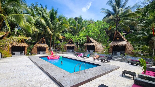A pool area with a clear blue pool, thatched-roof huts, and lounge chairs. - Flower Power Village (Ranong)