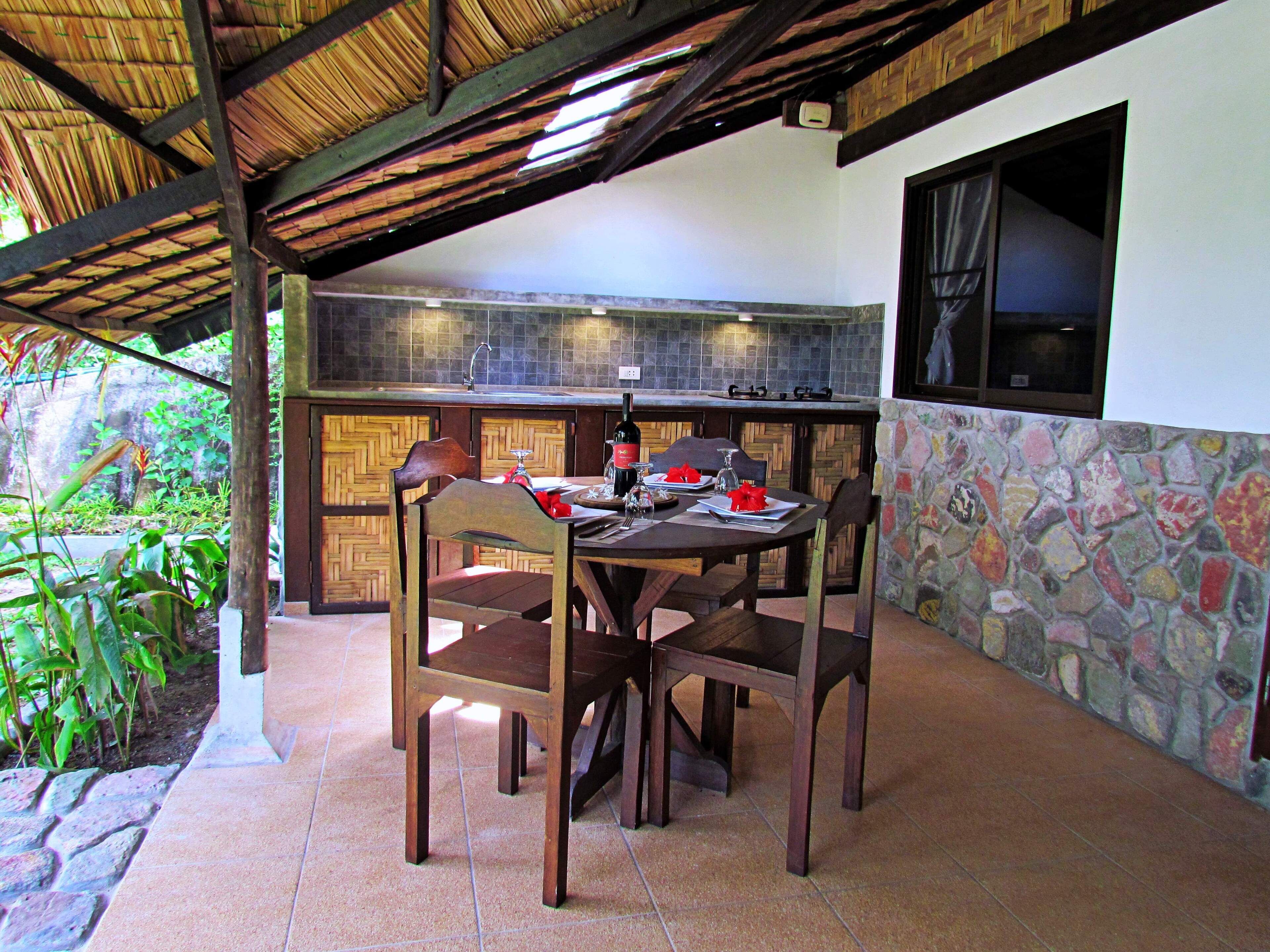An outdoor dining area with a thatched roof, stone walls, and a view of the surrounding greenery.