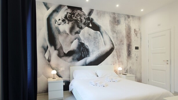 Un dormitorio con un mural grande en la pared, una cama con ropa de cama blanca, dos mesitas de noche con lámparas y una puerta blanca.