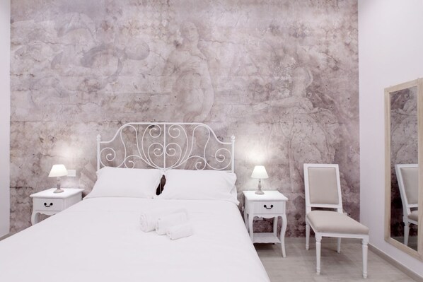 A bedroom with a marble wall, a white metal bed frame, two bedside tables with lamps, a chair, and a large mirror. - Suite Maggiore (Rome)