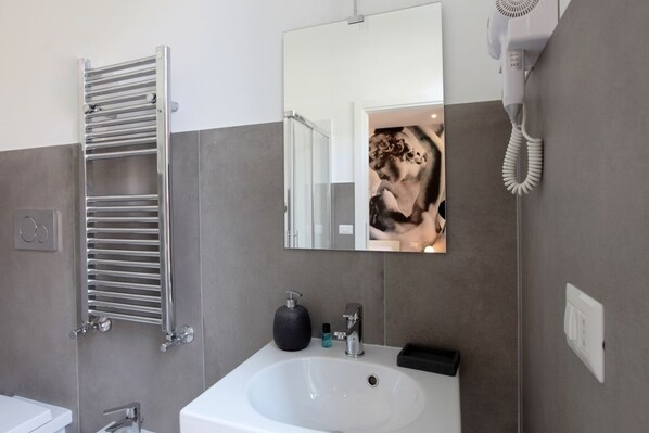 A modern bathroom with a white sink, a towel rack, a hairdryer, and a mirror reflecting a person. - Suite Maggiore (Rome)