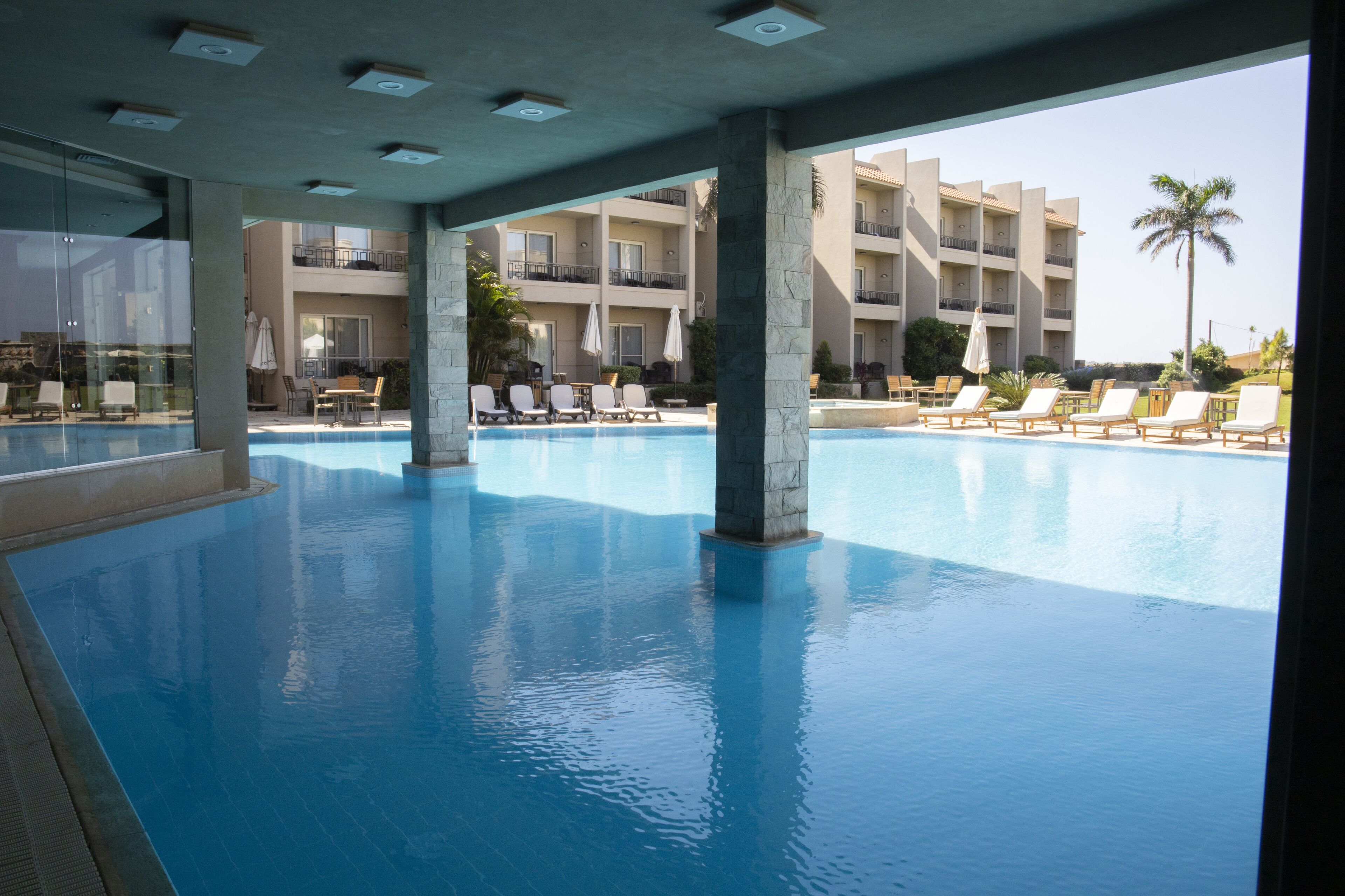 A large indoor swimming pool with lounge chairs and umbrellas, surrounded by a multi-story building with balconies.