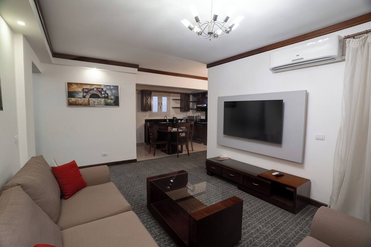 A living room with a sofa, TV, and entertainment unit. There is a dining area with a table and chairs in the background.