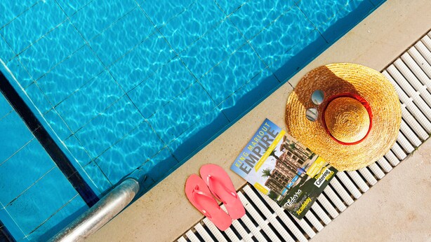 A poolside view with a tiled edge, a pair of pink flip-flops, a straw hat, and a magazine titled "King Mario Empire". - Panacea Suites Hotel (Borg el Arab)