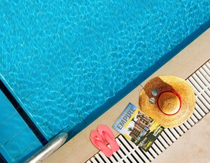 A poolside view with a tiled edge, a pair of pink flip-flops, a straw hat, and a magazine titled "King Mario Empire". - Panacea Suites Hotel (Borg el Arab)