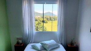 A bedroom with a bed, two nightstands, a window with curtains, and a view of greenery and hills.
