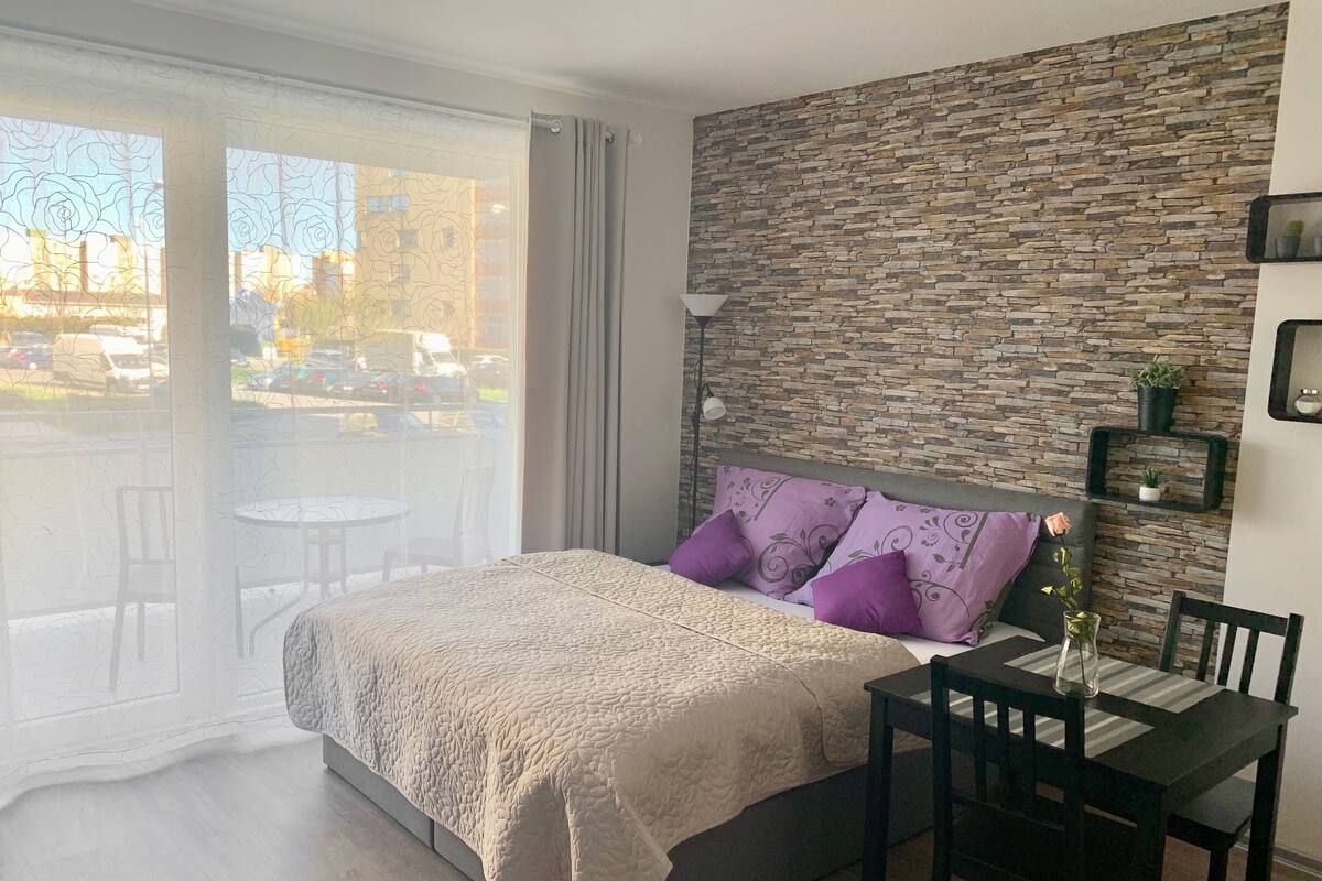 A bedroom with a bed, a dining table, chairs, a stone wall, and a view of the outside.