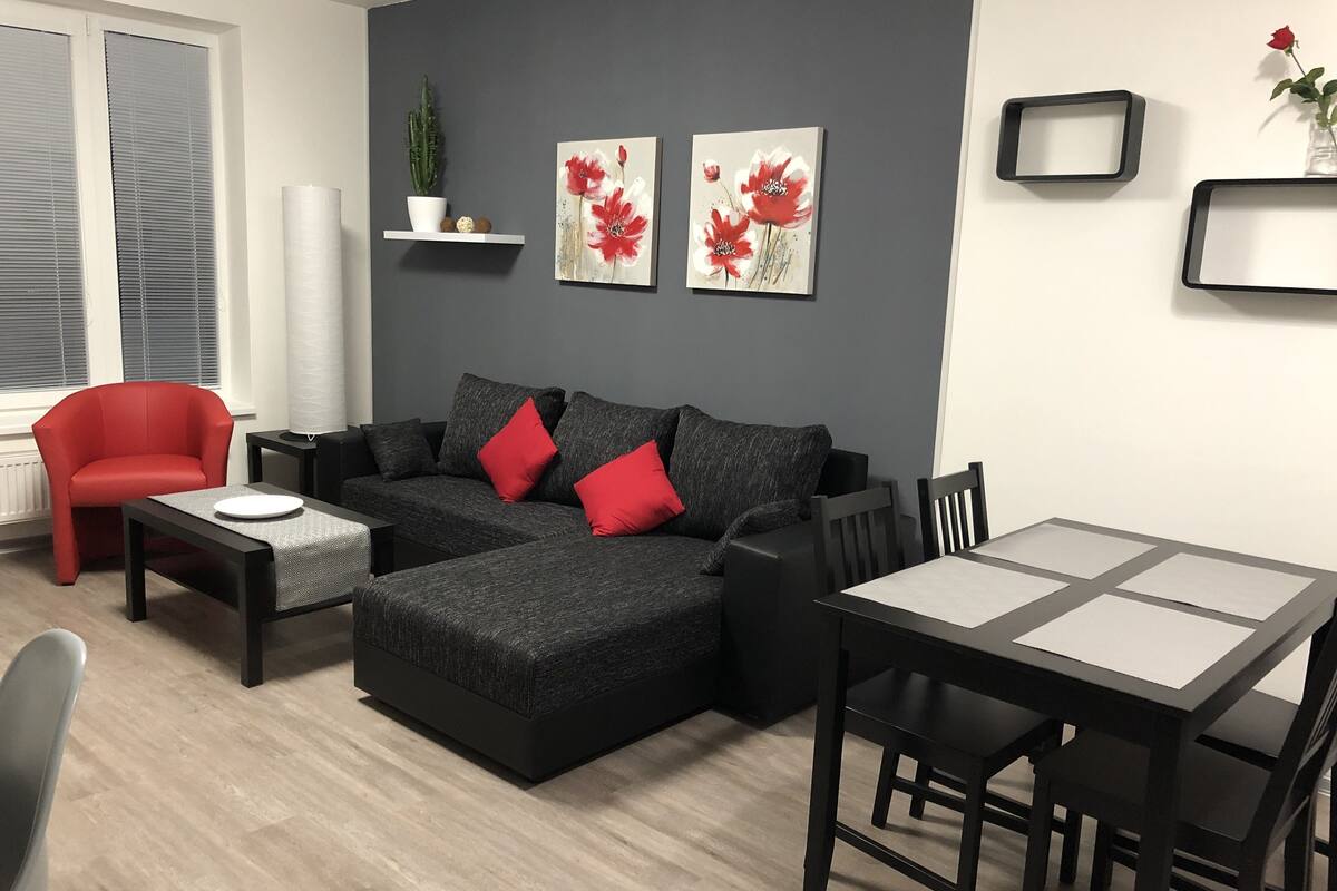 A modern living room with a grey sofa, red cushions, a red armchair, a small table, and a dining table with chairs. There are two wall-mounted paintings of red flowers and a cactus on a shelf.