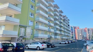 Un edificio residencial de varios pisos con balcones, un estacionamiento con coches y un cielo azul despejado.