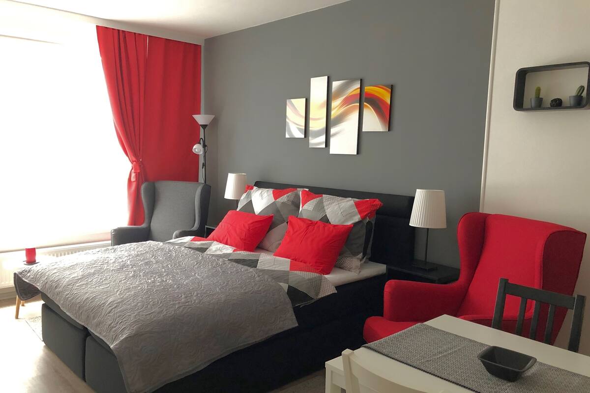 A modern hotel room with a bed, a desk, and a chair. There are two lamps on the desk and a red chair.