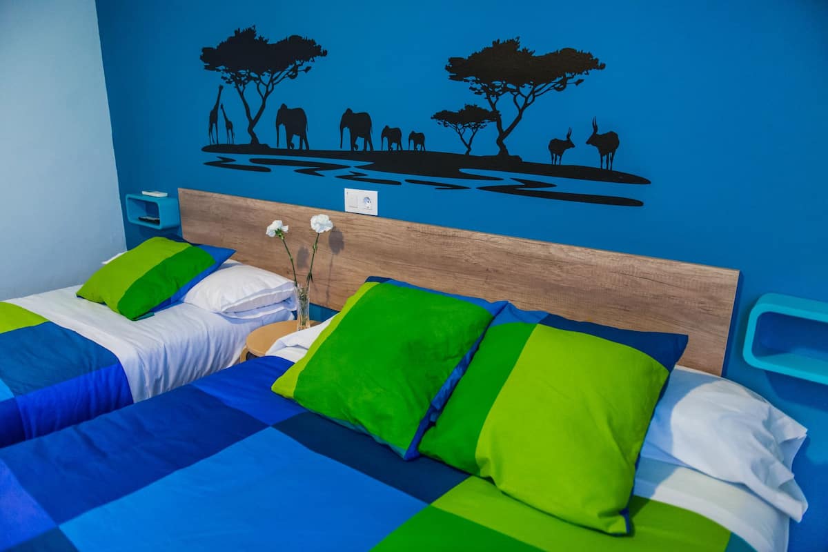 Une chambre avec deux lits, chacun agrémenté de draps bleus et verts, d’une tête de lit en bois et d’un motif décoratif mural représentant un paysage de savane.