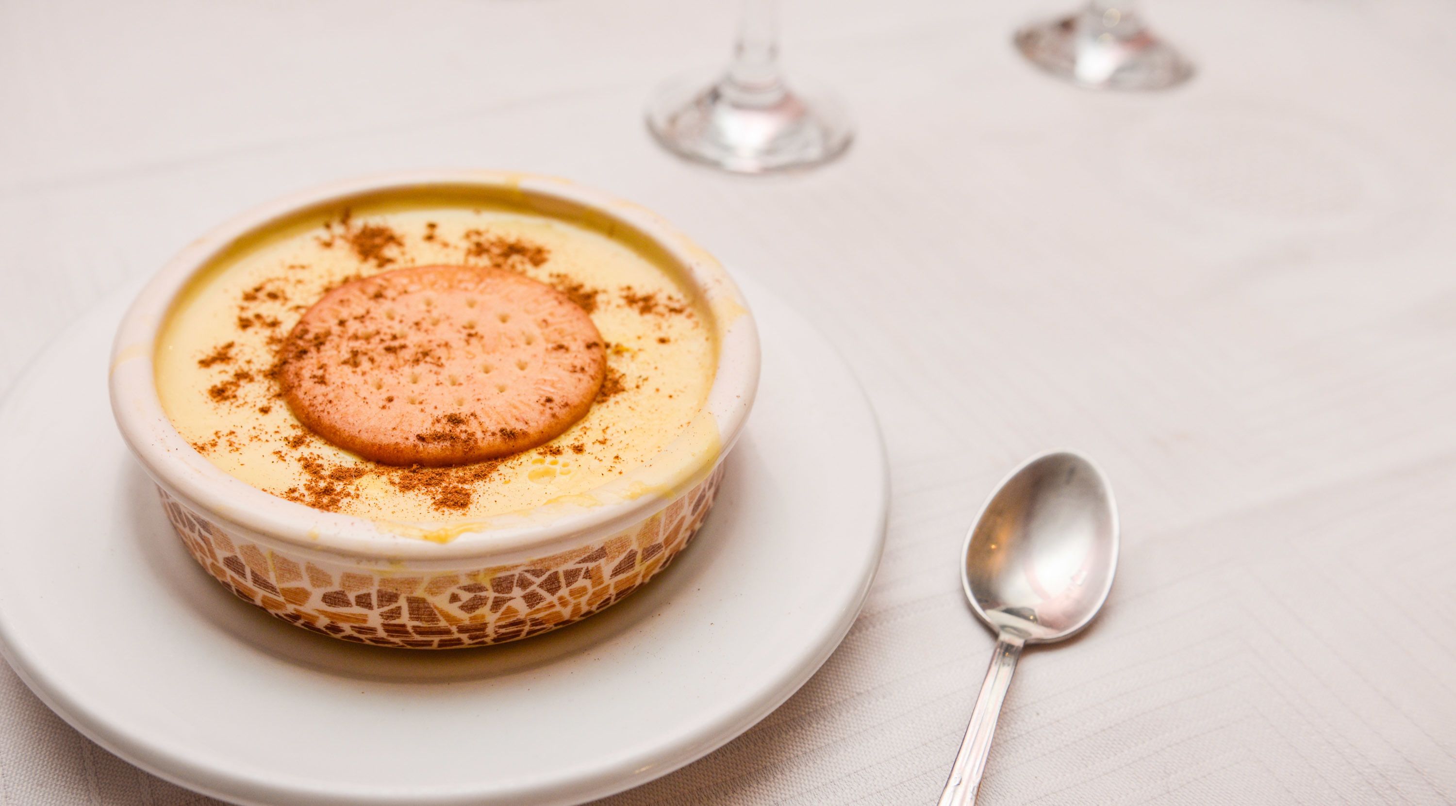 A dessert in a patterned bowl on a white plate with a spoon beside it.