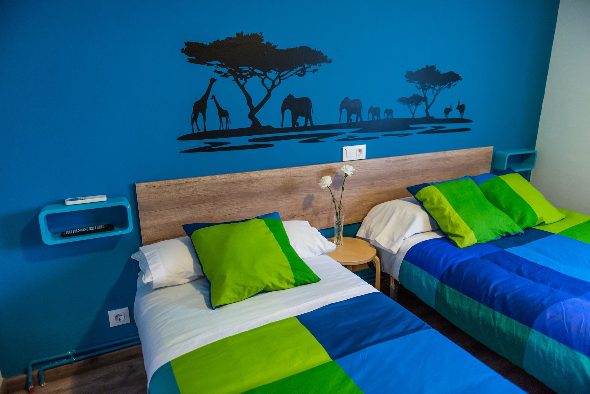Habitación con dos camas, cada una con una colcha azul y verde, cabeceros de madera y un mural de animales en la pared.