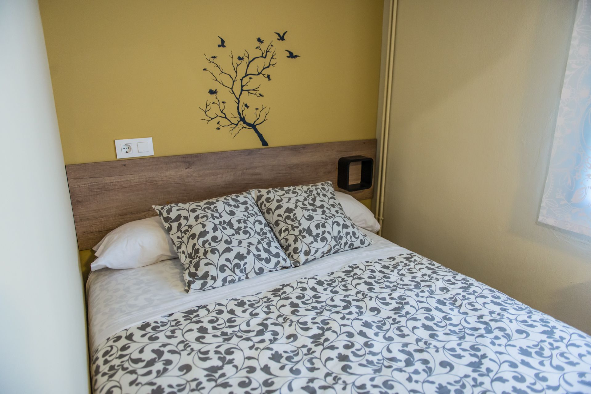 A bed with a patterned bedspread, a wooden headboard, and a wall decoration of a tree with birds.