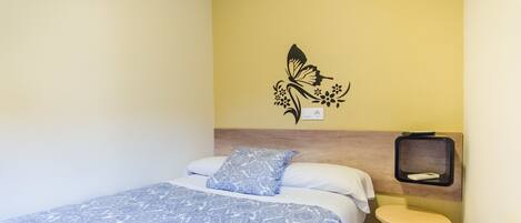 Una cama bien tendida con una colcha azul estampada, una almohada a juego y una mesita de noche fijada a la pared con un pequeño taburete.