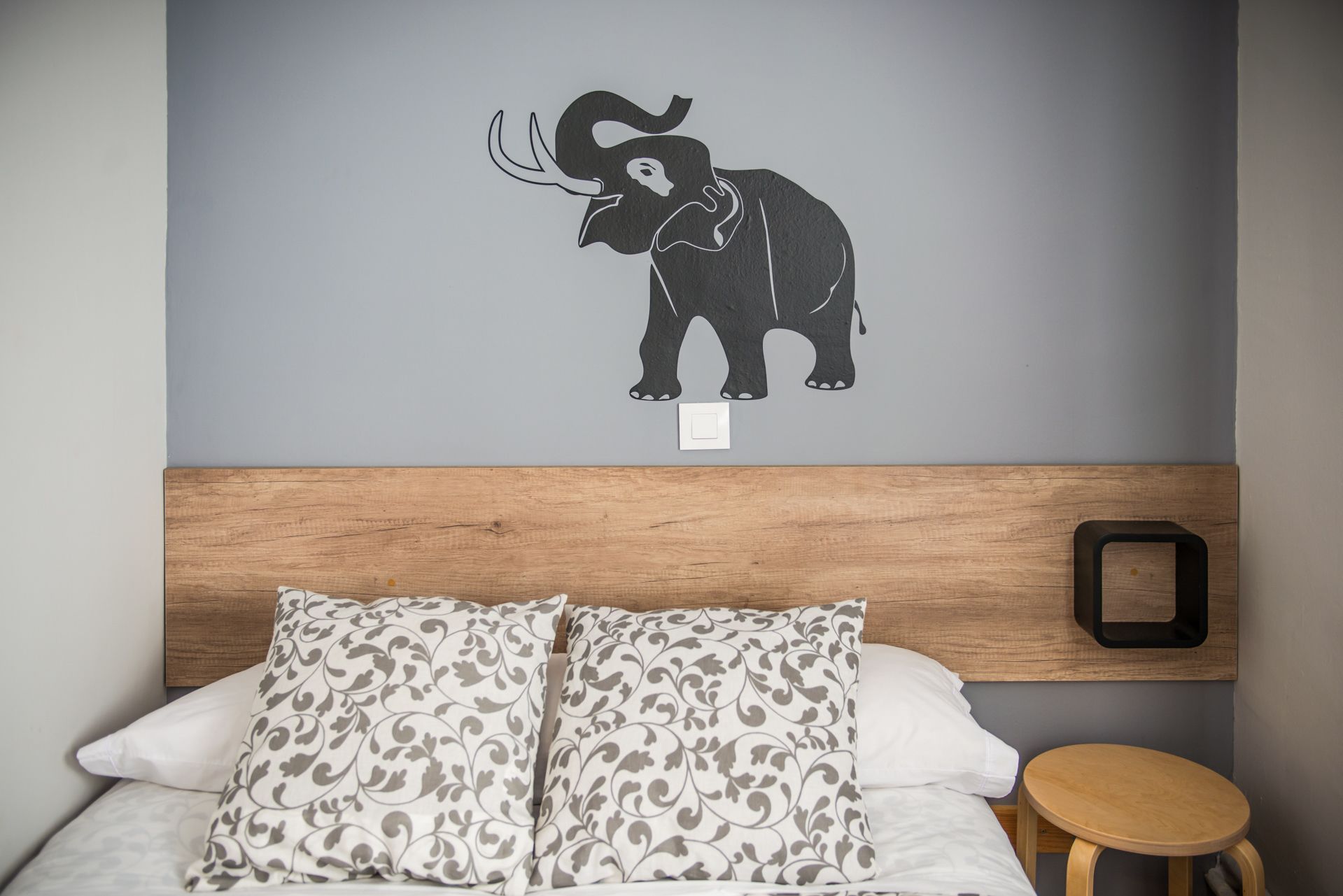 Een slaapkamer met een bed, twee kussens, een houten hoofdbord, een nachtkastje en een muursticker van een olifant.