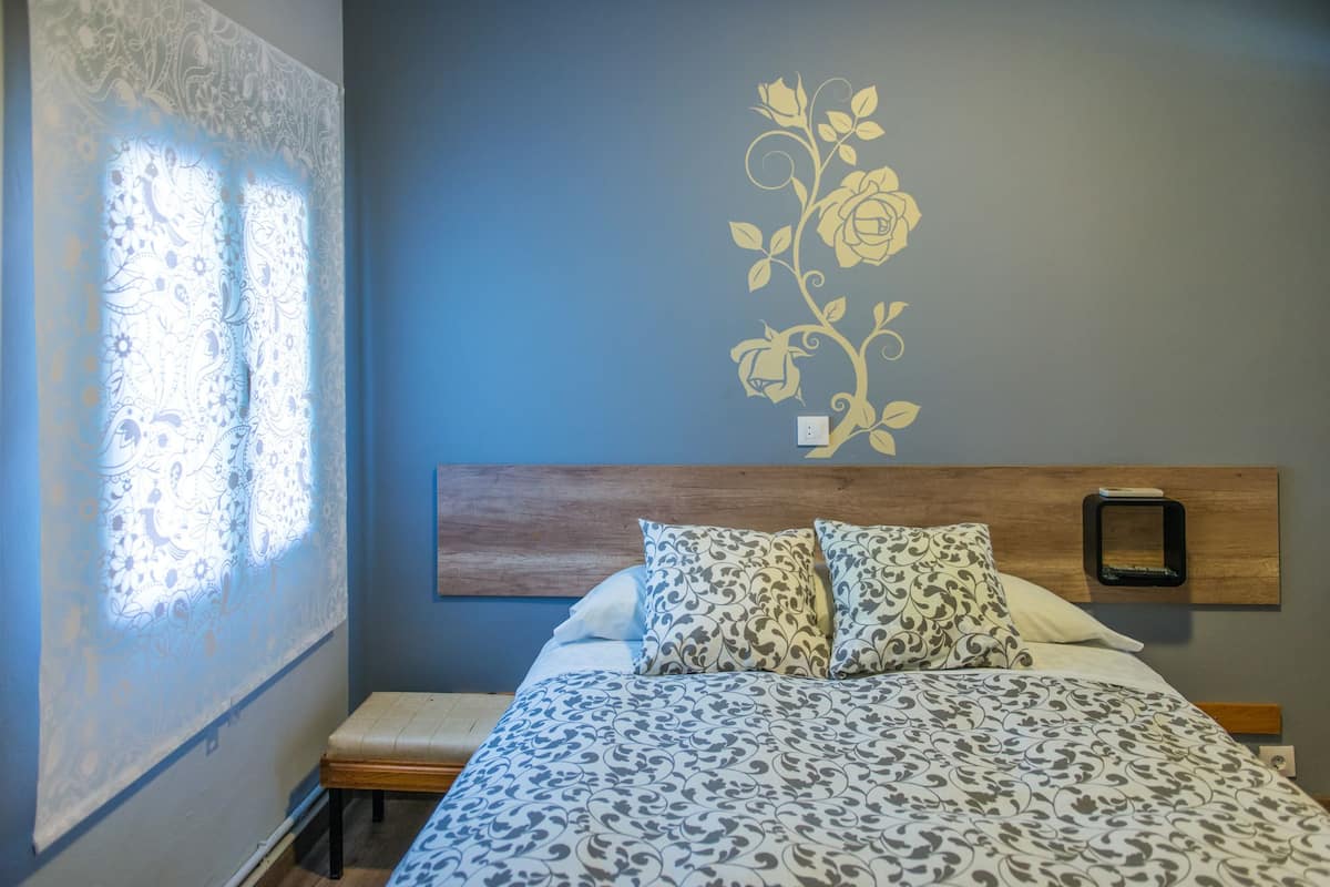 Une chambre à coucher comprenant un lit, deux oreillers, une tête de lit en bois, une table de chevet et un mur orné d’un motif floral.