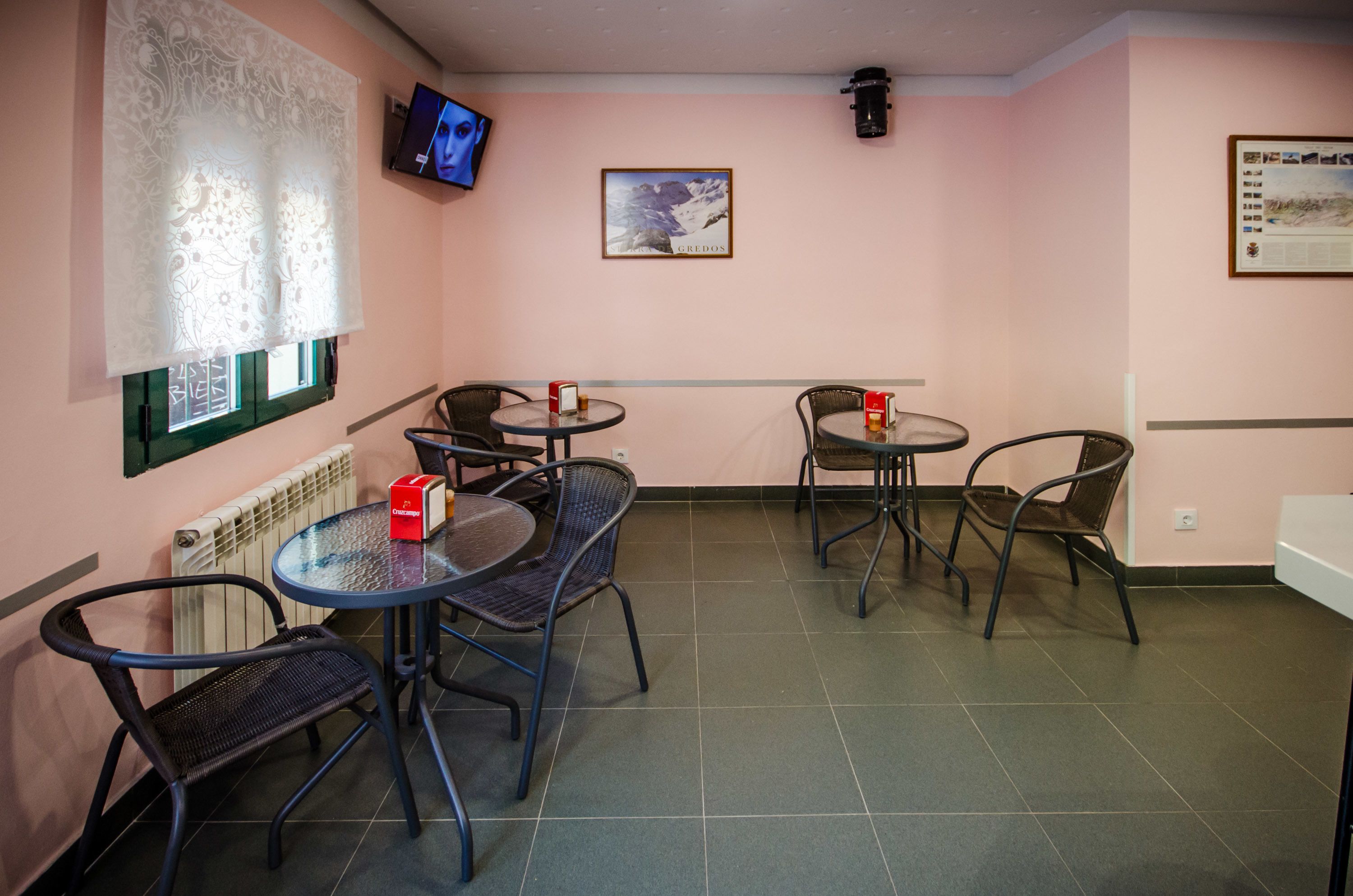 A room with a pink wall, a television mounted on the wall, and a framed picture. There are four tables and six chairs arranged in the room.