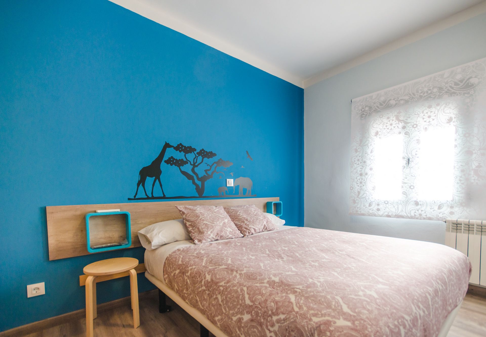 A bedroom with a bed, a nightstand, a small stool, a wall mural, and a window with a curtain.