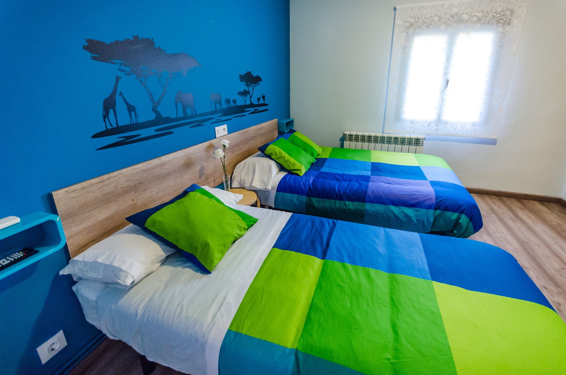 Un dormitorio con dos camas, cada una con colcha de cuadros azules y verdes, almohadas blancas y cabecero de madera. Se aprecia un mural en la pared con animales y árboles, una ventana con cortina blanca y una pequeña mesa con una silla azul.