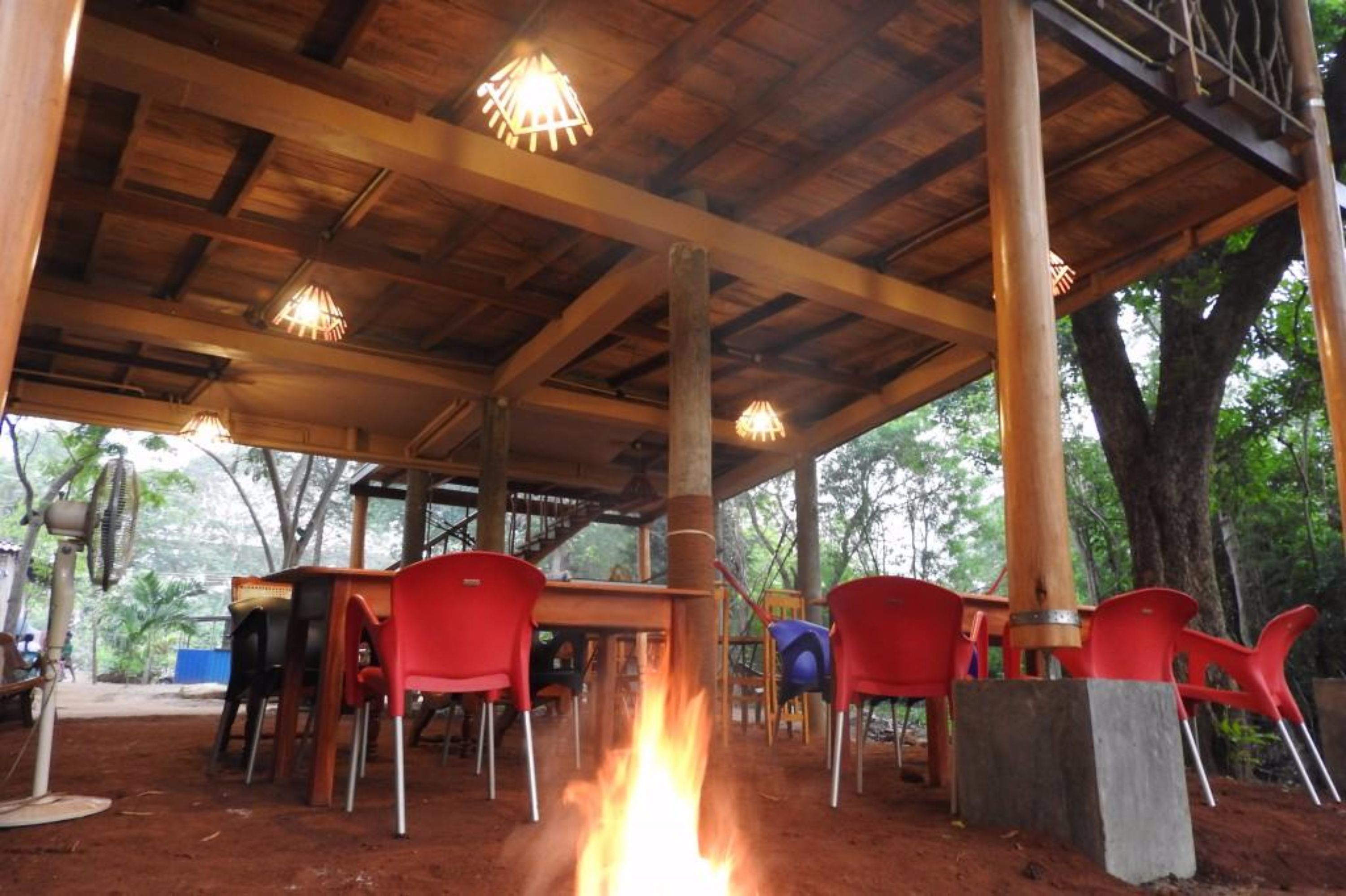 Un espacio de estar al aire libre, cubierto, con vigas de madera y luces colgantes, rodeado de árboles.