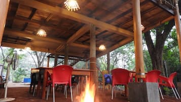 Un espacio de estar al aire libre, cubierto, con vigas de madera y luces colgantes, rodeado de árboles.