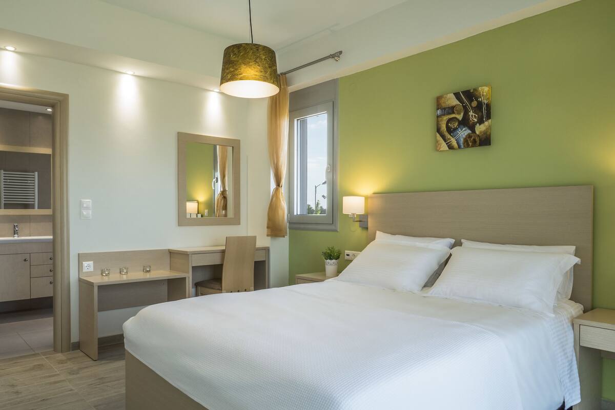 A modern hotel room with a large bed, a desk, a chair, a mirror, and a painting on the wall.