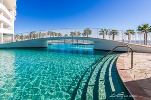 A clear blue pool with a bridge, surrounded by a white fence and palm trees.