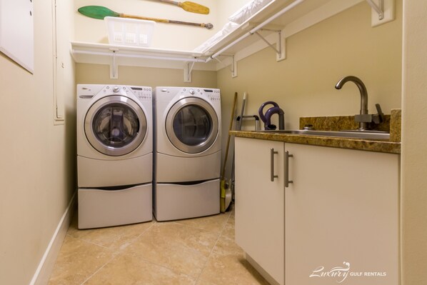 A laundry room with two front-loading washing machines, a sink with a faucet, and a countertop with a basket and cleaning tools.