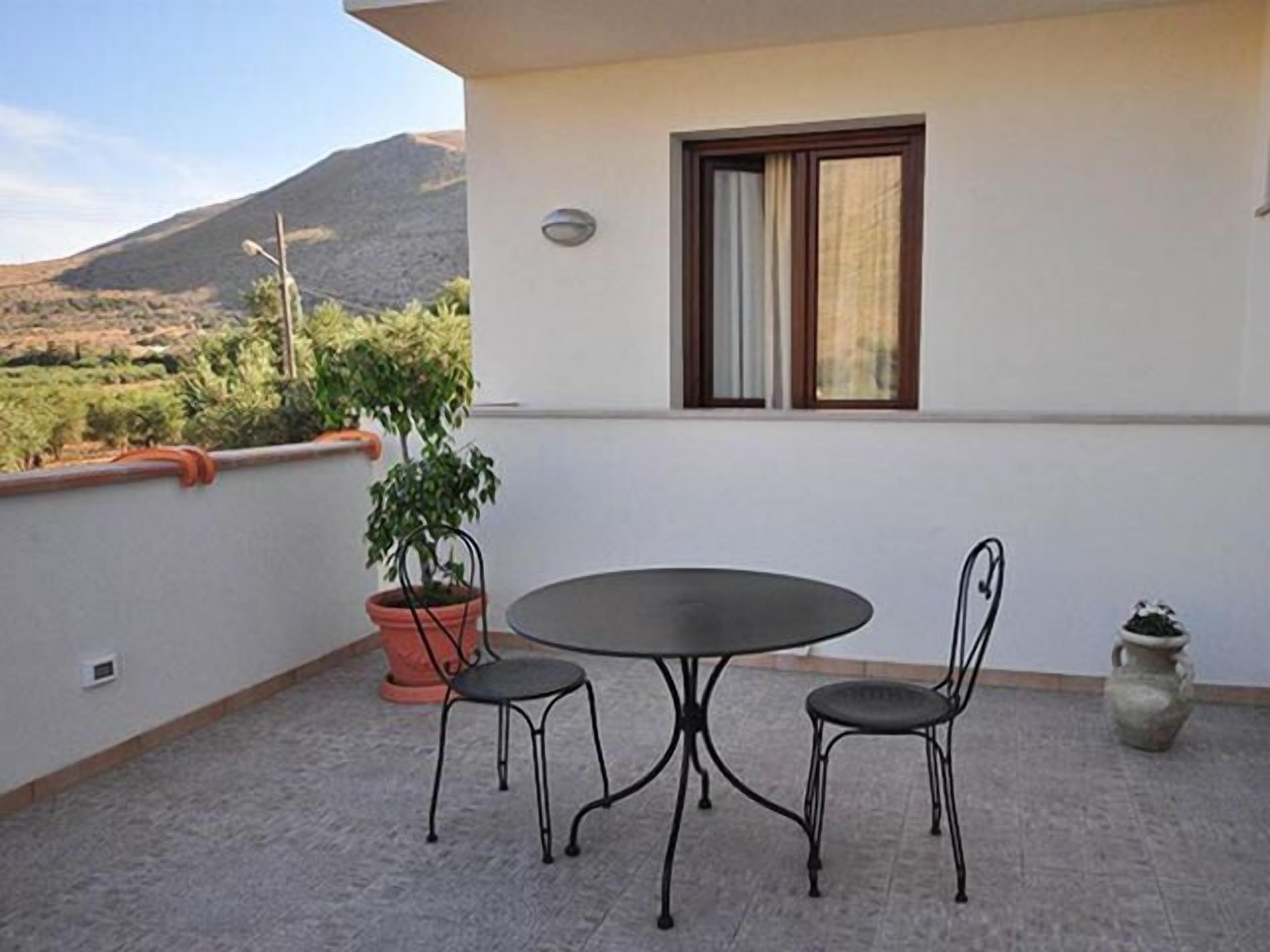 Uma varanda com mesa redonda e duas cadeiras, um vaso de planta e vista para uma paisagem montanhosa.