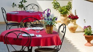 Un área de comedor al aire libre con mesas cubiertas de manteles rojos, sillas de metal negro y plantas en macetas.
