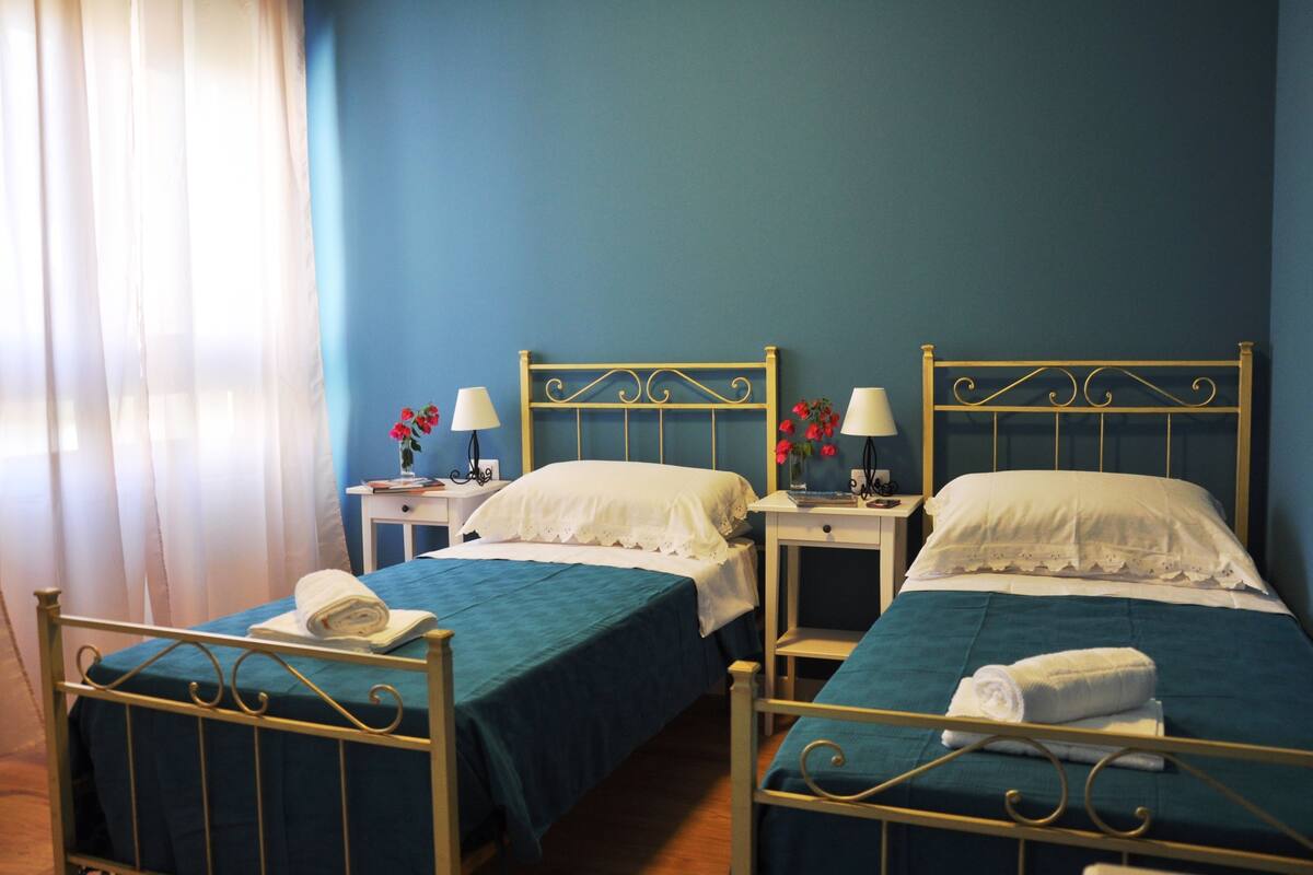 Una habitación de hotel con dos camas individuales, una mesita de noche con lámpara y una ventana con cortinas.