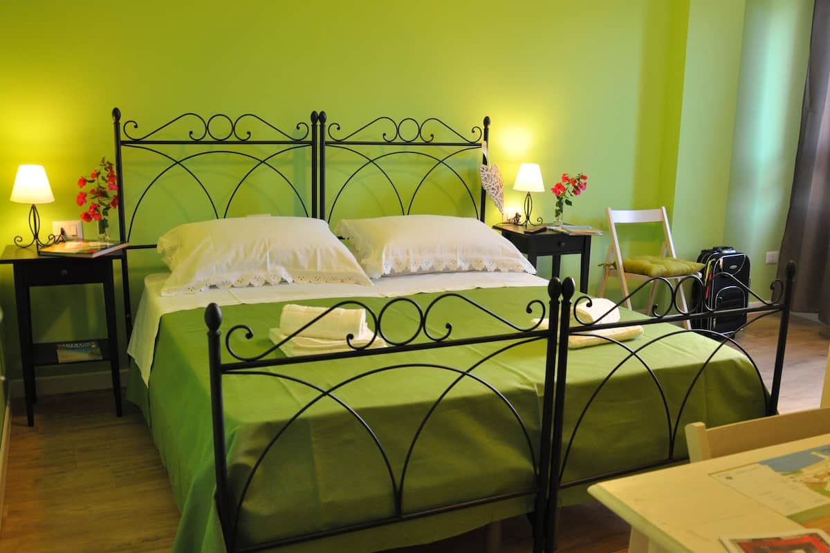 Una habitación con una cama verde, dos mesitas de noche con lámparas, una silla y una maleta.
