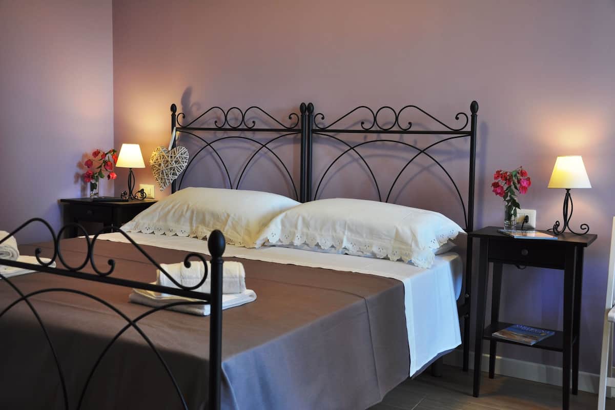 Una cama bien tendida con sábanas blancas, una mesita de noche con lámpara y un pequeño florero con flores.