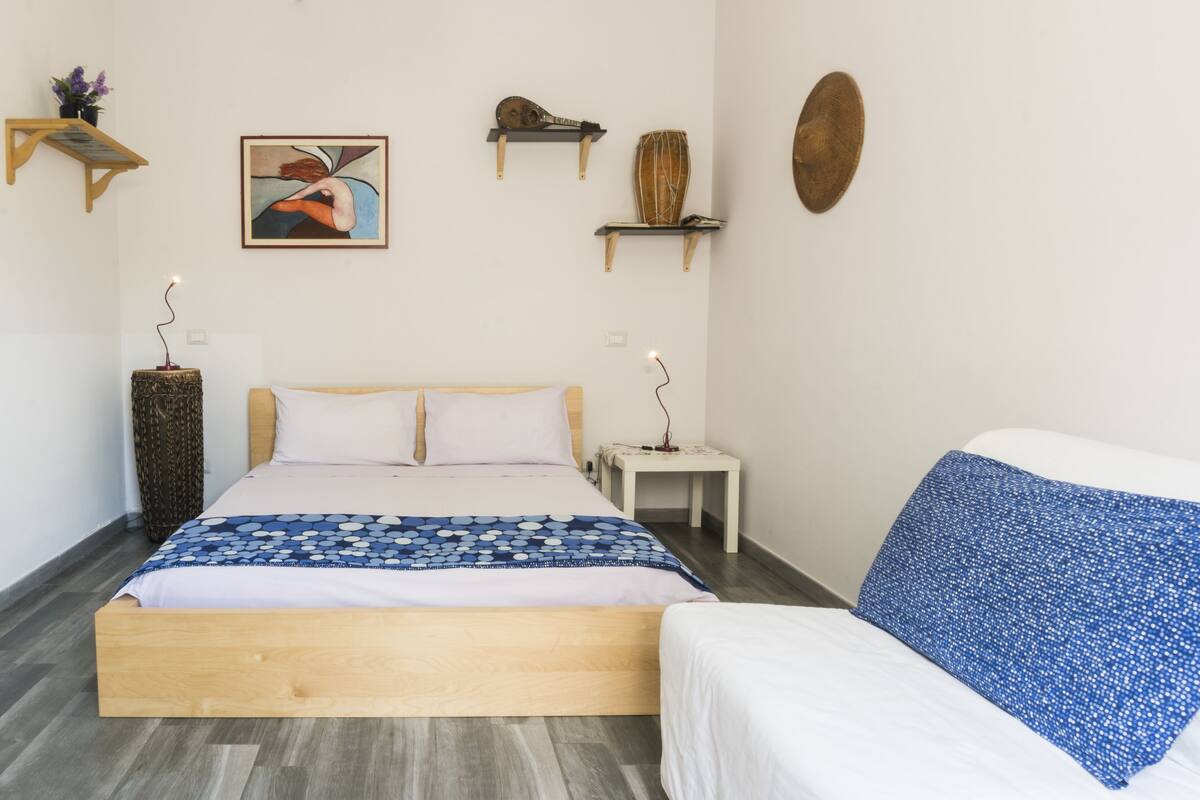 A bedroom with a wooden bed, a white bedside table, a small shelf with decorative items, and a wall-mounted shelf with a guitar and a vase.