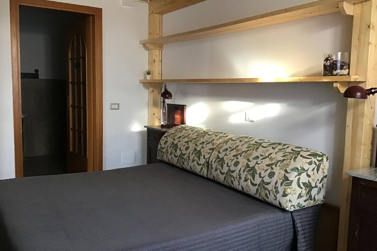 A bedroom with a bed, a nightstand, a lamp, a shelf with a vase, and a door.