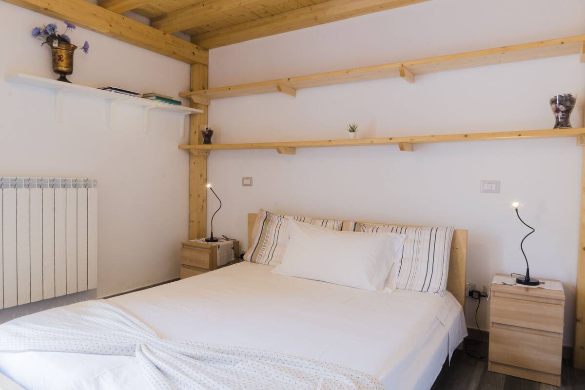 A bedroom with a wooden ceiling, a bed with white bedding, a nightstand with a lamp, and a radiator.