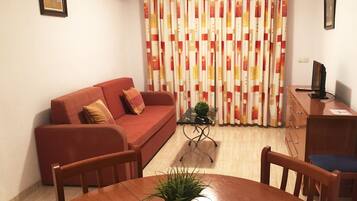 Una sala de estar con un sofá rojo, una mesita con una planta, un televisor sobre un mueble de madera y una cortina con estampado geométrico.