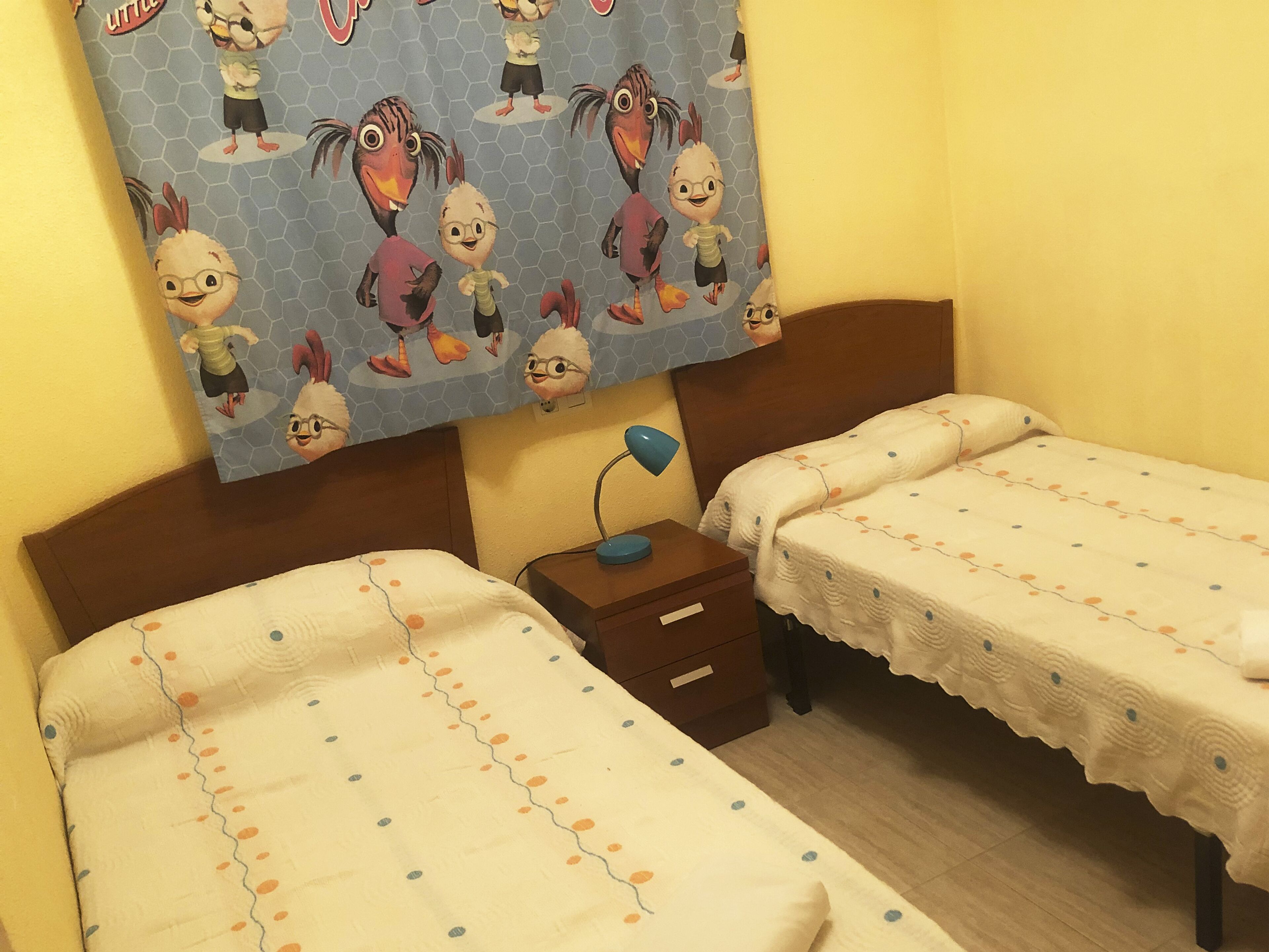 A room with two beds, each with a white bedspread featuring orange and blue polka dots. There is a nightstand with a lamp between the beds. Above the beds, there is a wall hanging with cartoon characters.