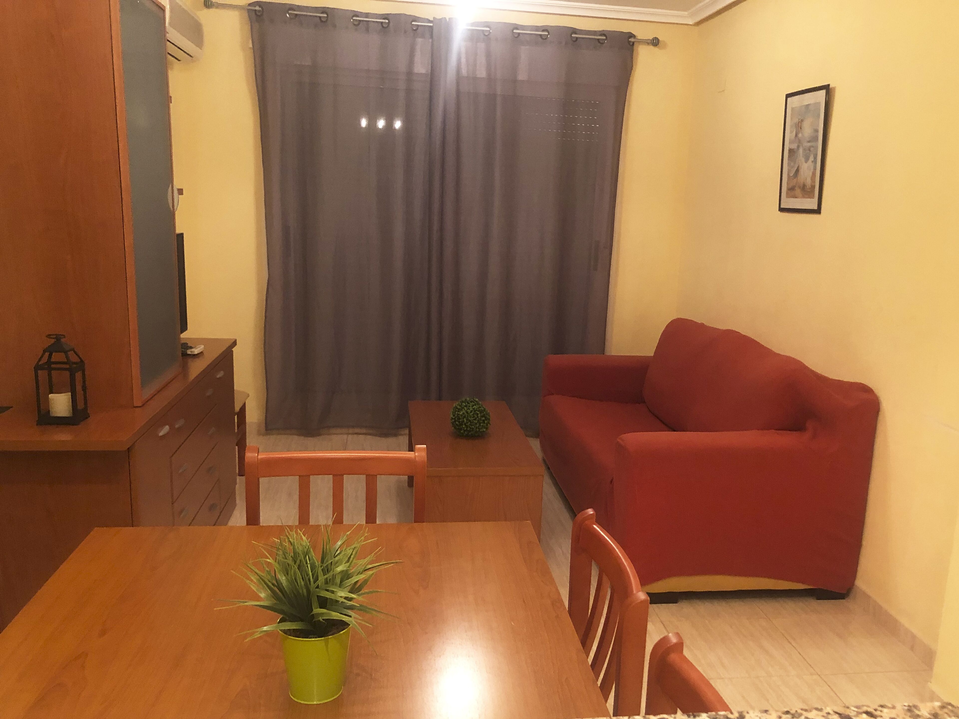 Una sala de estar con una mesa de madera, un sofá rojo, una planta verde y una cortina gris.